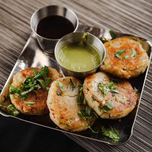 Four golden-brown Indian potato patties, garnished with cilantro, served with two dipping sauces on a metal tray.
