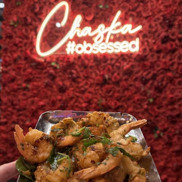Chaska Indian Restaurant's crispy fried shrimp with herbs and spices, against a red floral wall with a neon sign.